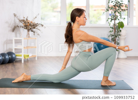Young woman yoga trainer performing crescent lunge pose on black rug 109933752