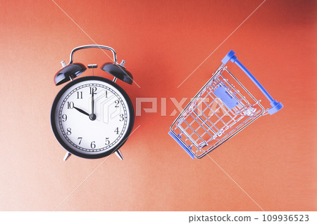An empty shopping cart and an alarm clock on a red scarlet background. Shopping, online shopping concept, time to buy concept, copy space, top view, 109936523