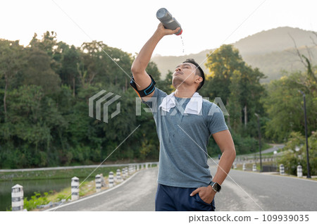 A thirsty, tired Asian man is drinking pouring water from a bottle on his face after a long run. 109939035