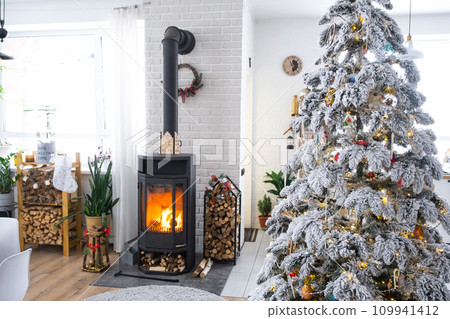 Festive white modern interior of house is decorated for Christmas and New Year in loft style with black stove, fireplace, Christmas tree. Potted plants, firewood in the woodpile 109941412