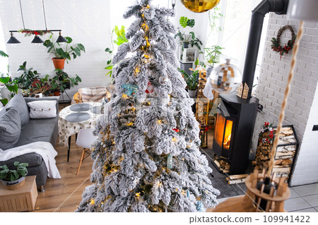 Festive white modern interior of house is decorated for Christmas and New Year in loft style with black stove, fireplace, Christmas tree. Potted plants, firewood in the woodpile Festive white modern interior of house is decorated for Christmas and New Year in loft style with black stove, fireplace, Christmas tree. Potted plants, firewood in the woodpile 109941422