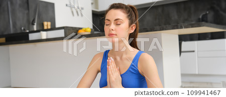 Young healthy beautiful woman in sports top and leggings practicing yoga at home, sitting in lotus position on yoga mat, meditating, smiling, relaxed, eyes closed, meditation concept Young healthy beautiful woman in sports top and leggings practicing yoga at home, sitting in lotus position on yoga mat, meditating, smiling, relaxed, eyes closed, meditation concept 109941467