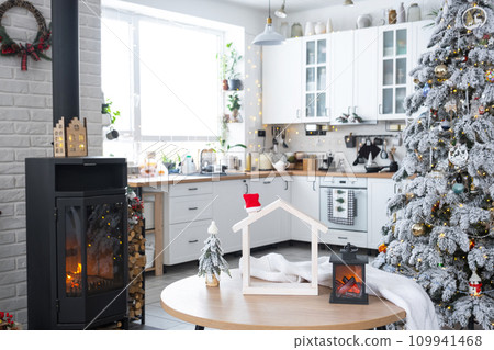 Christmas decor in white kitchen with burning stove fireplace, festive mess, village interior with a snowy Christmas tree. New Year, Christmas mood, cozy home. 109941468