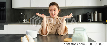 Attractive young cheerful girl baking at the kitchen, making dough, holding recipe book, having ideas 109941873