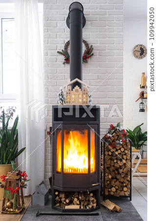 Metall black stove, burning hearth fireplace in white Festive interior of house is decorated for Christmas and New Year, Christmas tree. firewood in the woodpile, cozy and heating of home 109942050