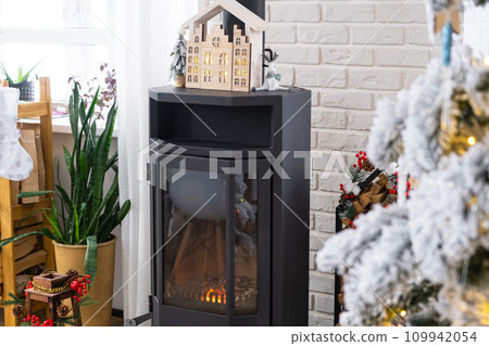 Metall black stove, burning hearth fireplace in white Festive interior of house is decorated for Christmas and New Year, Christmas tree. firewood in the woodpile, cozy and heating of home Metall black stove, burning hearth fireplace in white Festive interior of house is decorated for Christmas and New Year, Christmas tree. firewood in the woodpile, cozy and heating of home 109942054