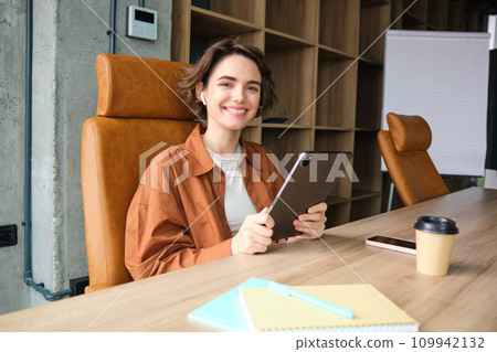 Portrait of young woman CEO, businesswoman in casual clothes, sitting in her office with digital tablet and cup of coffee, working on project, reading Portrait of young woman CEO, businesswoman in casual clothes, sitting in her office with digital tablet and cup of coffee, working on project, reading 109942132