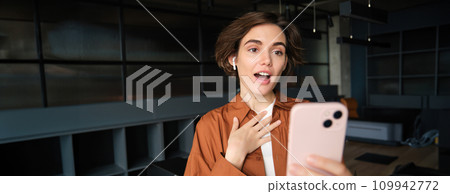 Portrait of working woman in office, looking surprised or shocked at smartphone screen 109942772