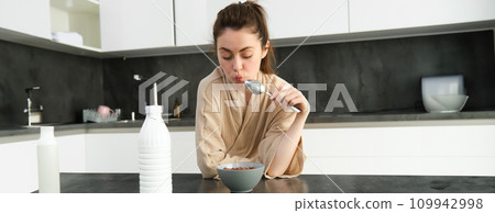 Portrait of young beautiful woman in bathrobe, eating cereals for breakfast, leans on kitchen worktop, looking at her morning meal 109942998