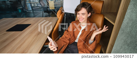 Business and young entrepreneurs concept. Young woman video chats, has online meeting, uses wireless headphones and smartphone app, smiling while talking Business and young entrepreneurs concept. Young woman video chats, has online meeting, uses wireless headphones and smartphone app, smiling while talking 109943017