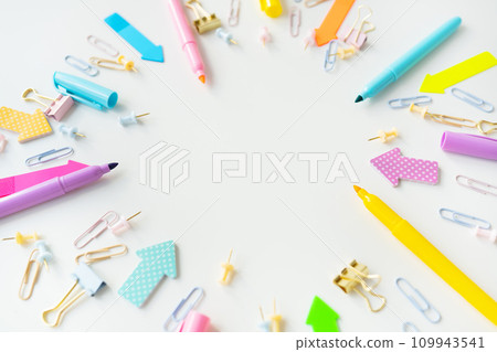 Colorful assortment of office supplies including pens, markers, paper clips and paper clips scattered on a white background. The overall mood is bright and creative, with space for writing. 109943541