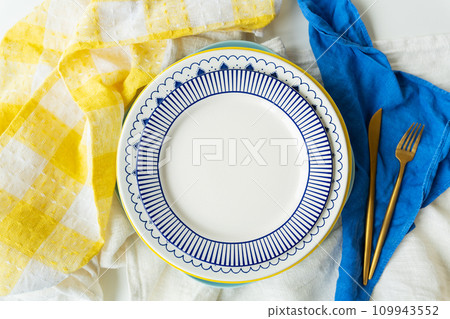 A white ceramic plate with an intricate blue pattern around the edge. The gold fork and knife are carefully placed on top of the dark blue cloth napkin. A white ceramic plate with an intricate blue pattern around the edge. The gold fork and knife are carefully placed on top of the dark blue cloth napkin. 109943552