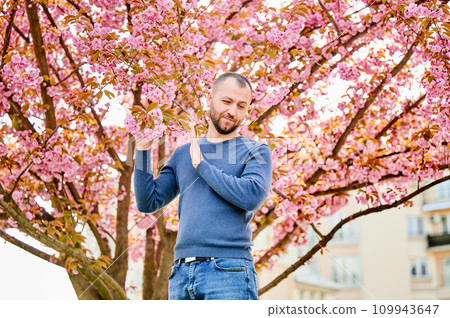 Man allergic suffering from seasonal allergy at spring in blossoming garden at springtime. Bearded man pushing away branch of blooming tree with flowers. Spring allergy concept Man allergic suffering from seasonal allergy at spring in blossoming garden at springtime. Bearded man pushing away branch of blooming tree with flowers. Spring allergy concept 109943647