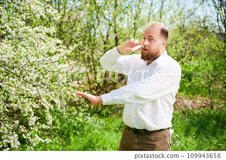 Man allergic using medical nasal drops, suffering from seasonal allergy at spring in blossoming garden. Handsome man treating runny nose in front of blooming tree outdoors. Spring allergy concept. Man allergic using medical nasal drops, suffering from seasonal allergy at spring in blossoming garden. Handsome man treating runny nose in front of blooming tree outdoors. Spring allergy concept. 109943658