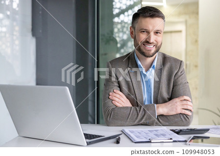 Portrait of successful financier accountant at workplace inside office, senior experienced businessman smiling looking at camera with crossed arms, working with papers sitting at table with laptop. 109948053