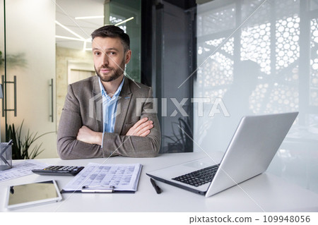 Serious and focused financier accountant on paper work inside office, mature man using calculator and laptop for calculating reports and summarizing accounts, businessman at work looking camera 109948056