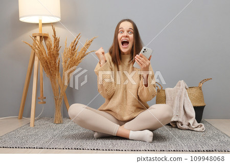 Checking social networks. Extremely happy Caucasian dark haired woman wearing casual clothing using phone with clenched fits and exclaiming happily sitting on floor in home interior 109948068