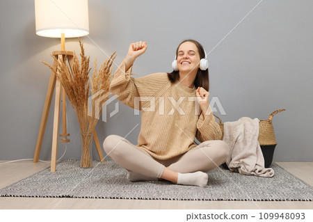 Overjoyed cheerful woman with brown hair wearing beige casual clothing and headphones hearing favorite song on radio dancing being in good mood sitting on floor in light room 109948093