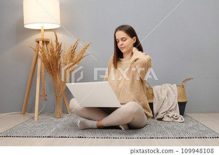 Freelance flexibility. Remote work. Tired overworked brown haired woman wearing beige sweater working on computer having pain in neck while sitting on floor in home interior 109948108