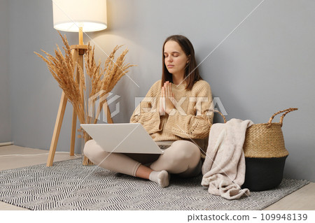Online break. Networking workspace professional. Calm brown haired woman wearing beige sweater working on computer practicing yoga having break meditating while sitting on floor in home interior 109948139