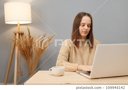 Remote worker using laptop. Online occupation. Concentrated beautiful brown haired woman wearing beige sweater working on computer while sitting at table against lamp and dry flowers 109948142