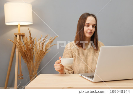Distance work options. Remote workspace. Calm brown haired woman wearing beige sweater working on computer and drinking coffee while sitting at table against lamp and dry flowers 109948144