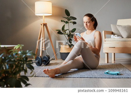 Sport and fitness online. Yoga with smartphone. Athletic workout at home. Fitness app for training. Sporty woman wearing beige tracksuit sitting on floor using mobile cell phone Sport and fitness online. Yoga with smartphone. Athletic workout at home. Fitness app for training. Sporty woman wearing beige tracksuit sitting on floor using mobile cell phone 109948161