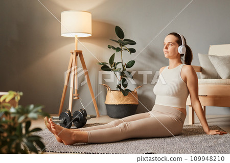 Calm sporty woman sitting on the floor practicing yoga wear tip and leggings meditating in living room listening meditation in headphones looking away with concentrated face 109948210