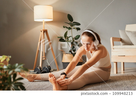Gymnastics in the house. Fit lifestyle at home. Exercising for health indoors. Pilates for relaxation. Young woman in headphones in sportswear doing fitness stretching exercises at home 109948213