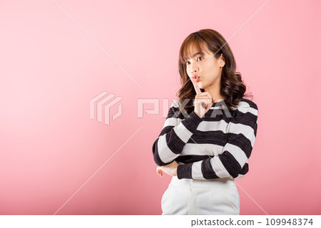 A woman in a studio shot on pink background signals quiet with a finger on her lips, inviting secrecy and mystery. 109948374