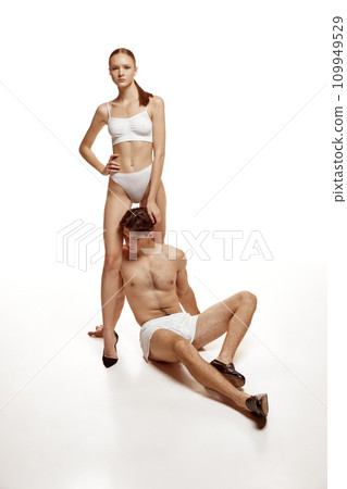 Portrait of beautiful couple, man sitting on floor and woman stands under him and put her hand on on males head against white background. 109949529