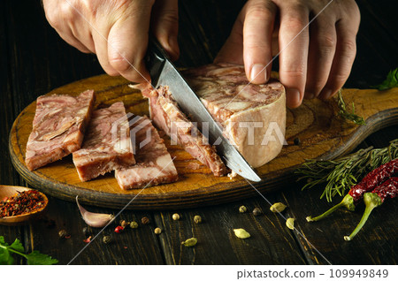 The cook cuts the brawn with a knife on the kitchen board before preparing sandwiches. Idea for a delicious snack with headcheese and aromatic spices. The cook cuts the brawn with a knife on the kitchen board before preparing sandwiches. Idea for a delicious snack with headcheese and aromatic spices. 109949849