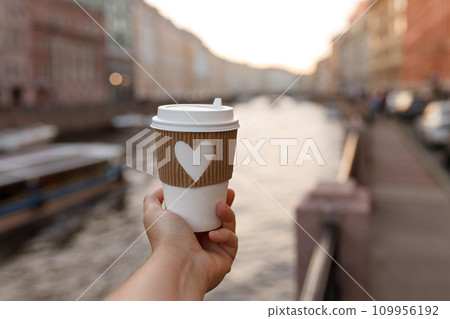 paper cup of drink in woman's palm on background of Fontanka river in St. Petersburg in summer day 109956192