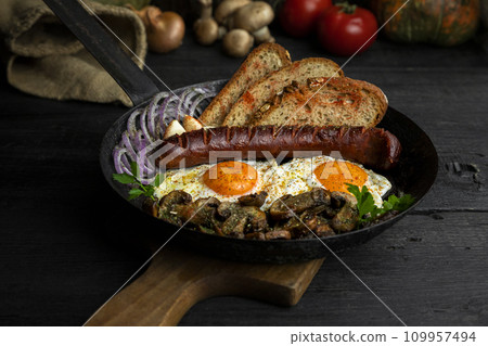 Fried eggs and sausage with mushrooms served in cast iron pan in the kitchen Fried eggs and sausage with mushrooms served in cast iron pan in the kitchen 109957494