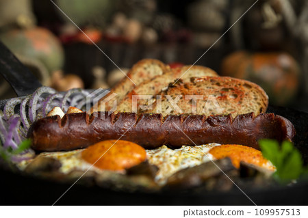 Sausage and fried eggs with toasted bread, mushrooms and onions served in cast iron dish. Selective focus on the sausage 109957513