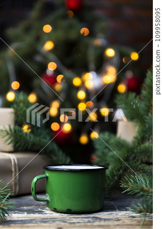 Coffee in a tin mug on the rustic table with wrapped gifts, Christmas tree lights and spruce branches in the background 109958095
