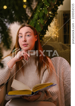 Hello winter. smiling modern woman in white sweater siting on chair and reading a book in room decorated for celebrating the new year and christmas festive mood 109958535