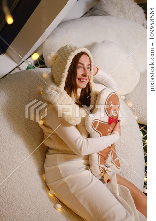 Merry young fun woman wearing white warm sweater hat posing hold ginger cookie man pillow studio portrait. Happy New Year celebration holiday concept 109958563