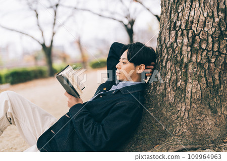 A man leaning against a tree and reading a book 109964963