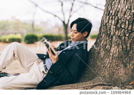 A man leaning against a tree and reading a book 109964969