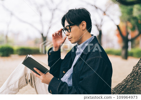 A man leaning against a tree and reading a book 109964973