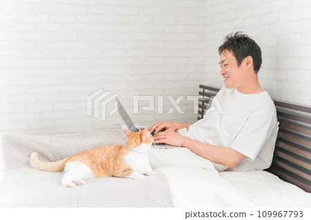 A man who telework/remote work with a cat/pet in a bedroom/bedroom at home A man who telework/remote work with a cat/pet in a bedroom/bedroom at home 109967793