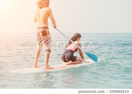 Sea woman and man on sup. Silhouette of happy young woman and man, surfing on SUP board, confident paddling through water surface. Idyllic sunset. Active lifestyle at sea or river. 109968810