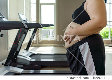 A plus-sized woman placing her hand on her belly, standing confidently in a well-equipped gymnasium. Side view A plus-sized woman placing her hand on her belly, standing confidently in a well-equipped gymnasium. Side view 109971109