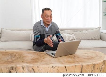 Old man relaxing comfortably on a sofa, immersed in operating a laptop. Evident focus and concentration in a cozy domestic backdrop. 109971113