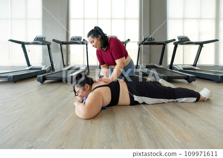 A plus-size lady is resting on the ground by a treadmill, looking tired but okay. A friend massages her back to help relieve pain. 109971611