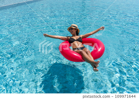 A lady enjoy a sunny day, floating on a bright inflatable ring in a clear blue swimming pool. A lady enjoy a sunny day, floating on a bright inflatable ring in a clear blue swimming pool. 109971644