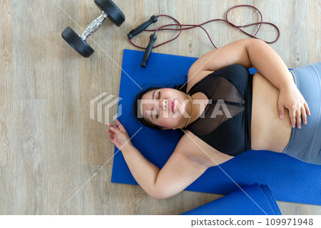 A plus-size woman lies exhausted on a blue mat, taking a breather after a vigorous workout session with dumbbell and jump rope. Top view A plus-size woman lies exhausted on a blue mat, taking a breather after a vigorous workout session with dumbbell and jump rope. Top view 109971948