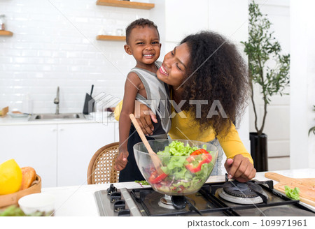 A smiling woman and her young son are making a fresh salad together in a cozy, well-equipped kitchen. 109971961