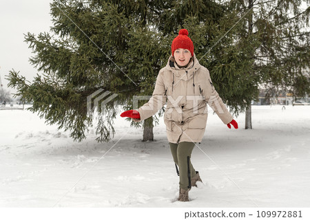 Pretty young woman in youth winter clothes is fooling around in snow near fir trees in park. Frosty winter day. 109972881